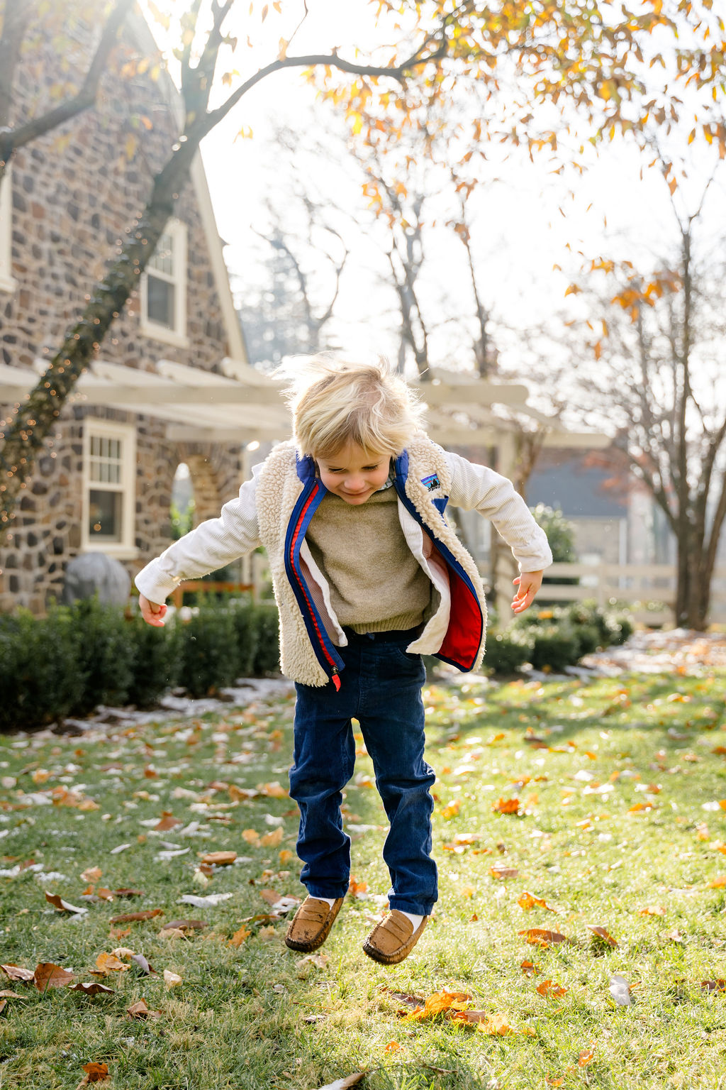 A toddler jumping in fall leaves outside a stone house